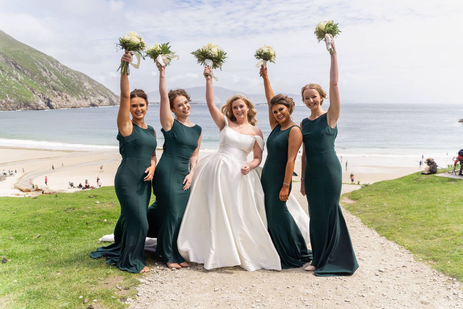 Bride in a white gown poses with four bridesmaids in emerald green dresses, all raising bouquets, on a grassy coastal beach wedding setting.