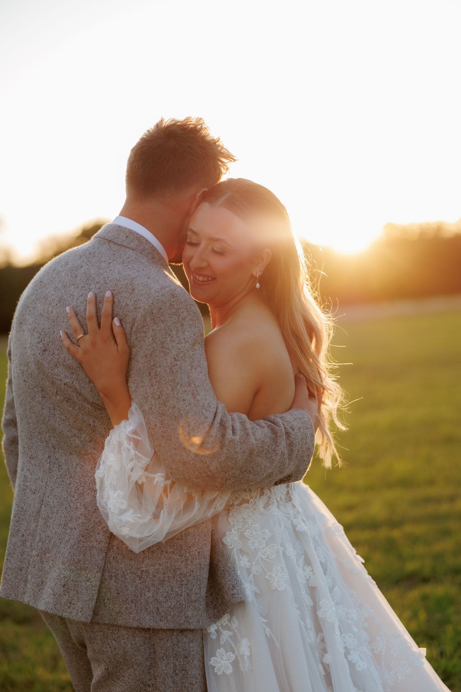 Bride in a lace wedding gown embraces her groom in a gray tweed suit in a grassy field at golden hour sunset.