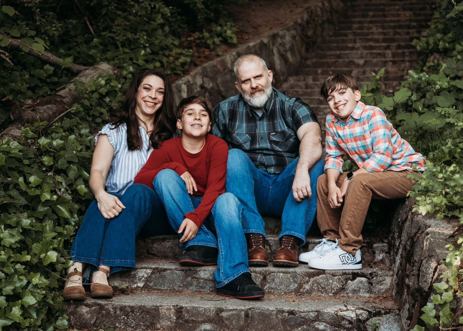 A smiling family of four sits together on mossy outdoor stone steps surrounded by green ivy, posing for a casual portrait.