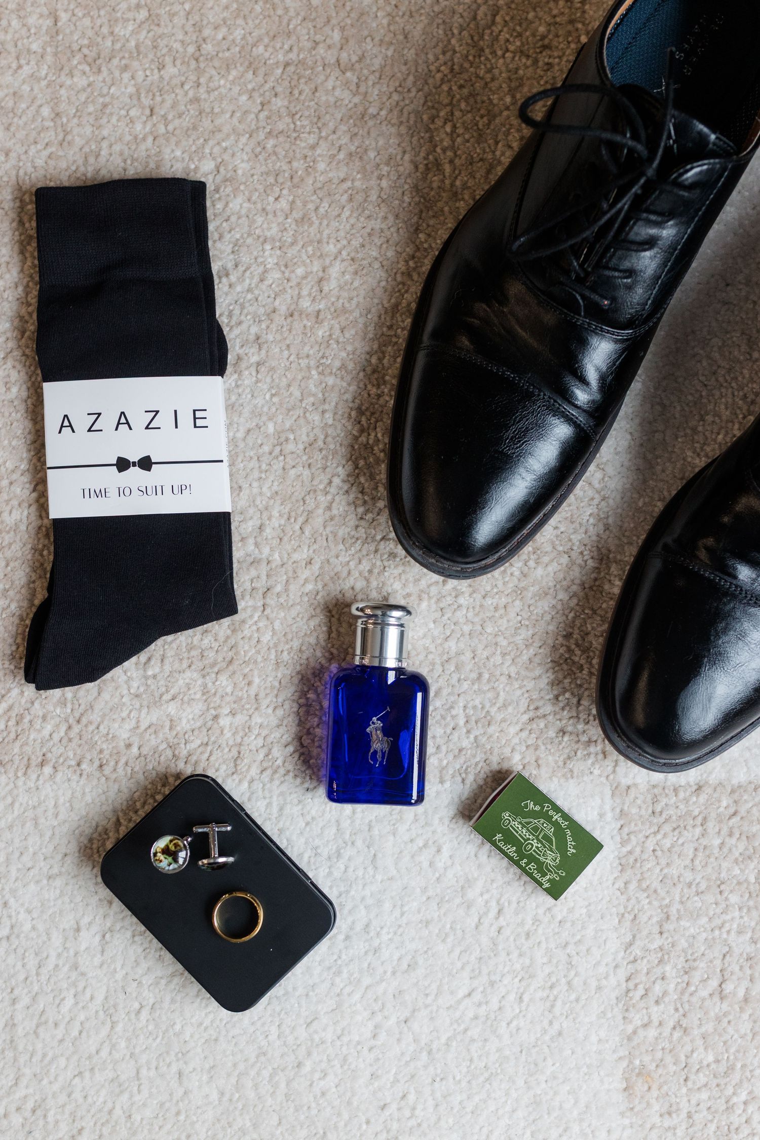 Groomsman wedding day flat lay with black dress shoes, Azazie socks, blue cologne bottle, cufflinks, a ring and a matchbook on a beige rug.