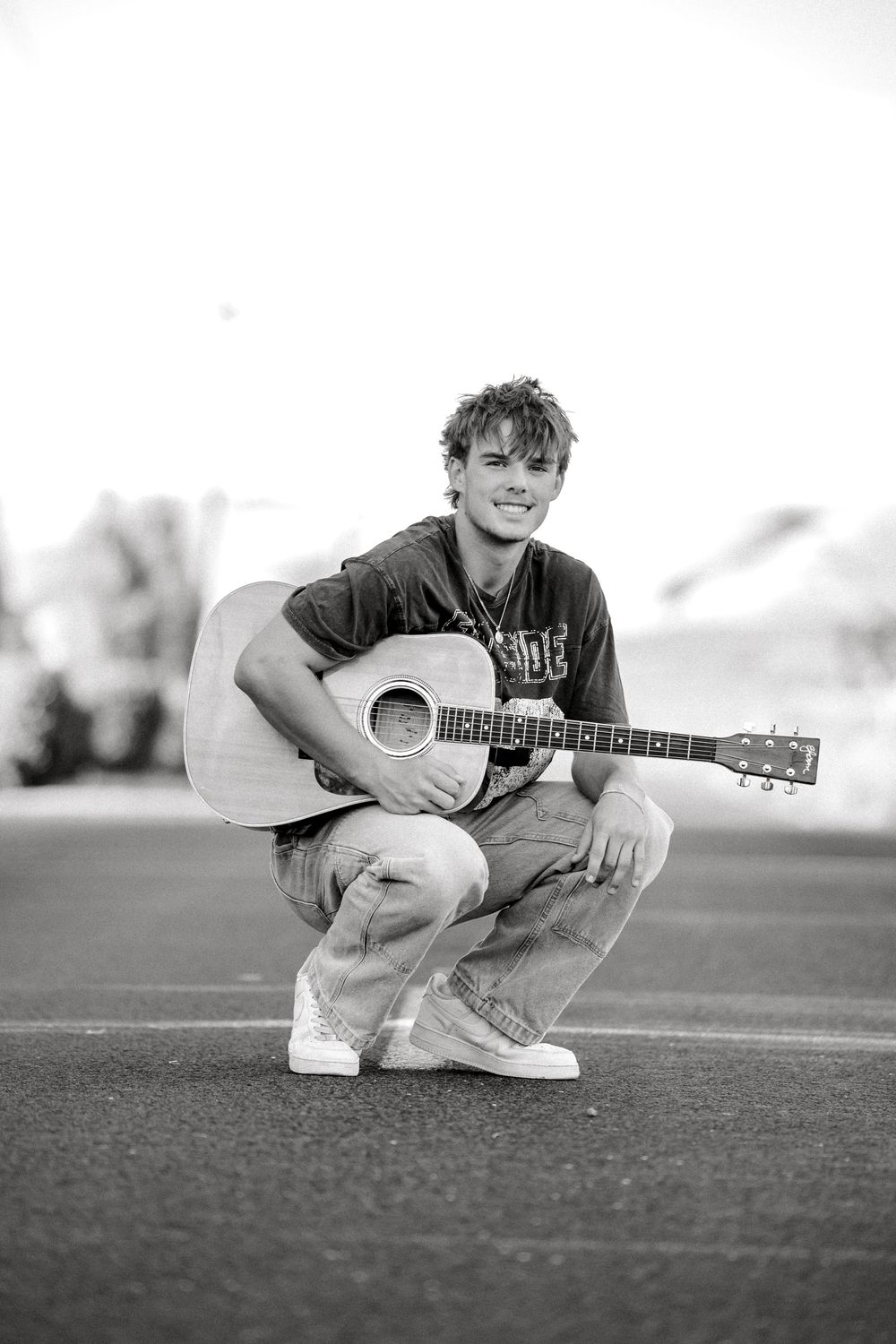 Young man crouching outdoors holding an acoustic guitar, smiling in a black and white photo.