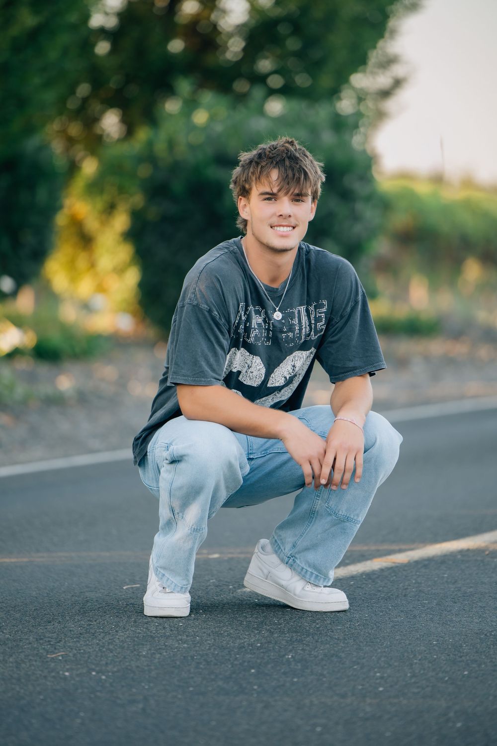 Young man crouching on a road in light jeans and gray graphic tee, smiling with green trees in background.