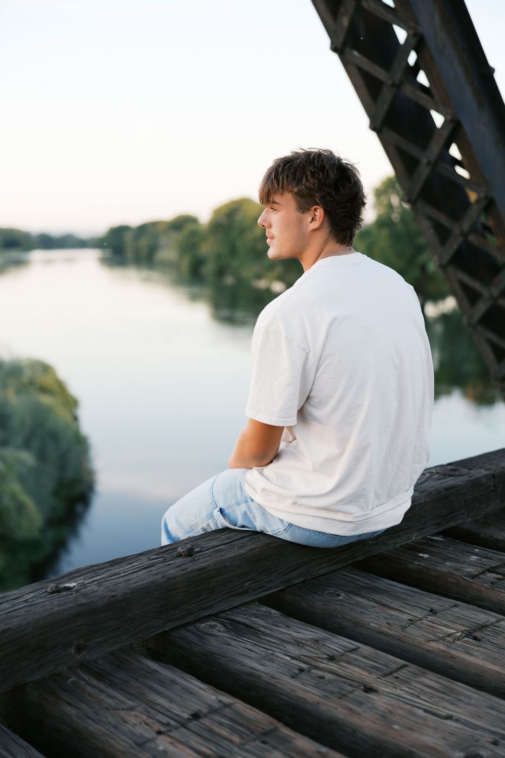 Young man in white t-shirt sitting on wooden bridge overlooking calm river surrounded by green trees at dusk.