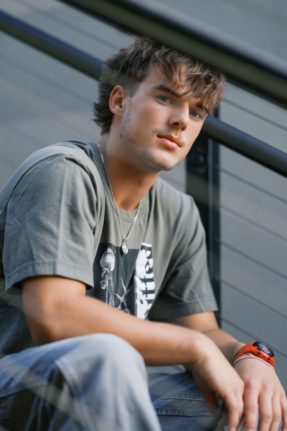 Young man in gray graphic tee and jeans sits on outdoor steps, wearing a red watch, with a relaxed pose.