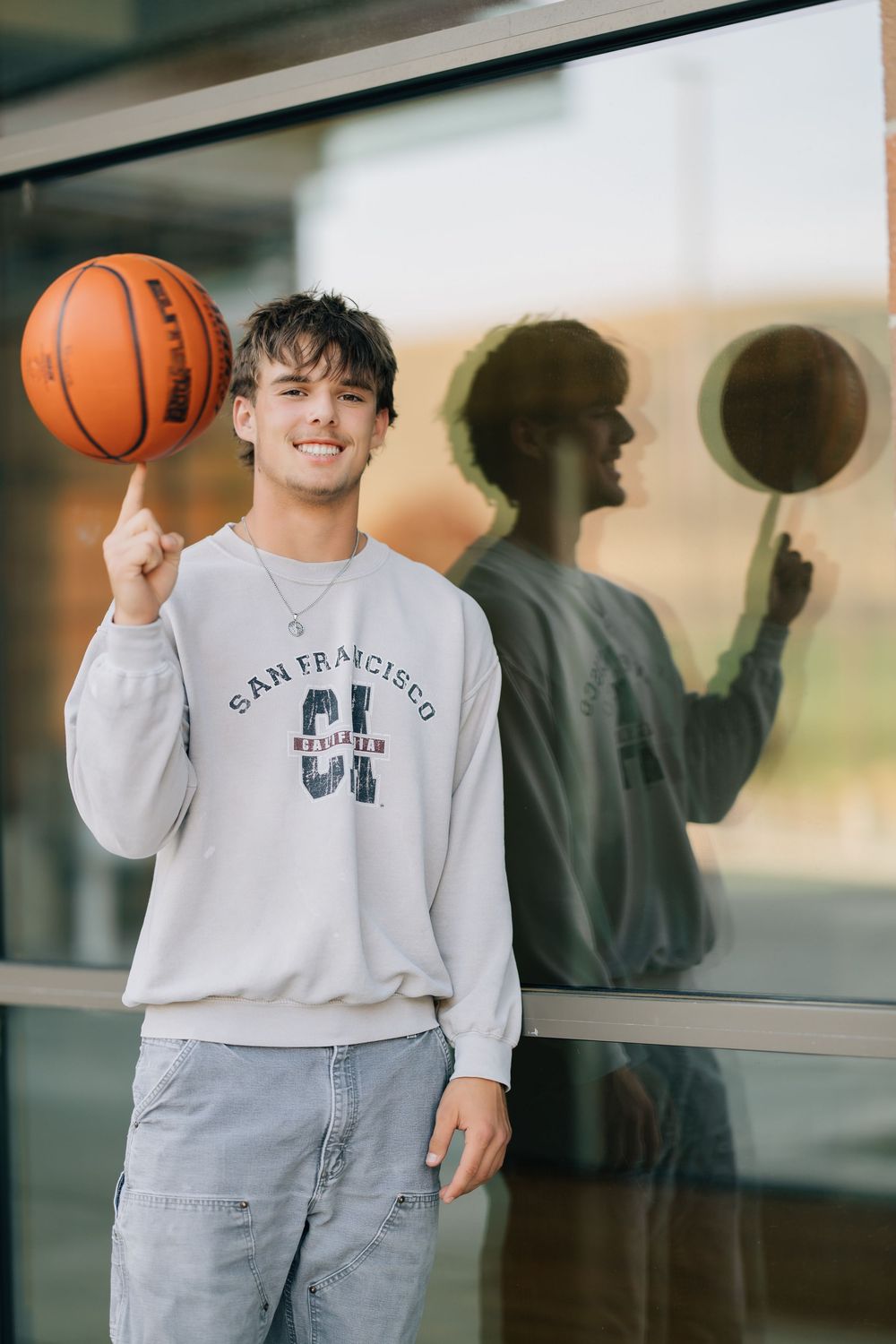 Young man in San Francisco sweatshirt spinning basketball on finger, smiling outdoors.
