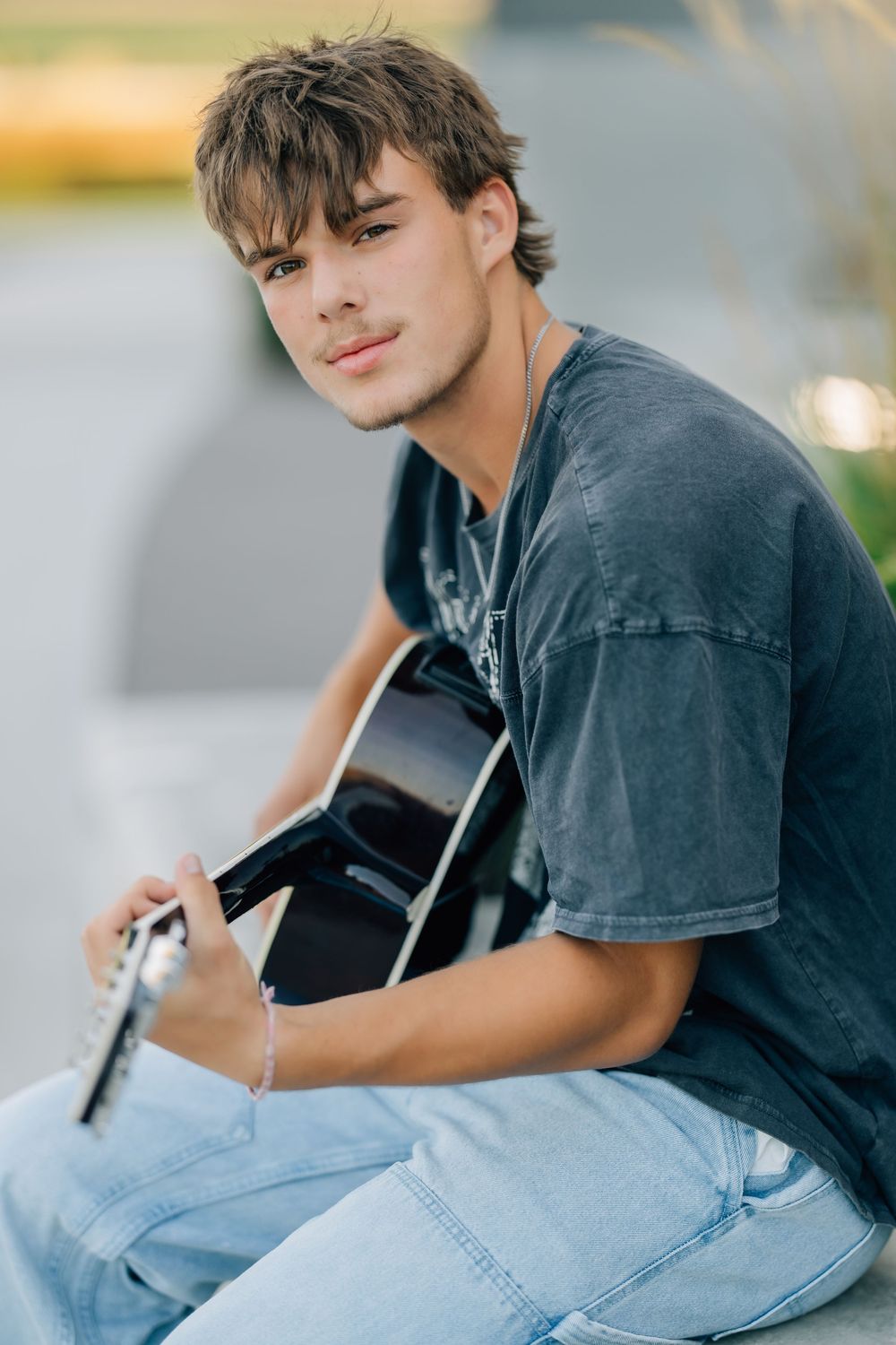 Young man in gray t-shirt and jeans playing acoustic guitar outdoors, looking at camera with relaxed expression.