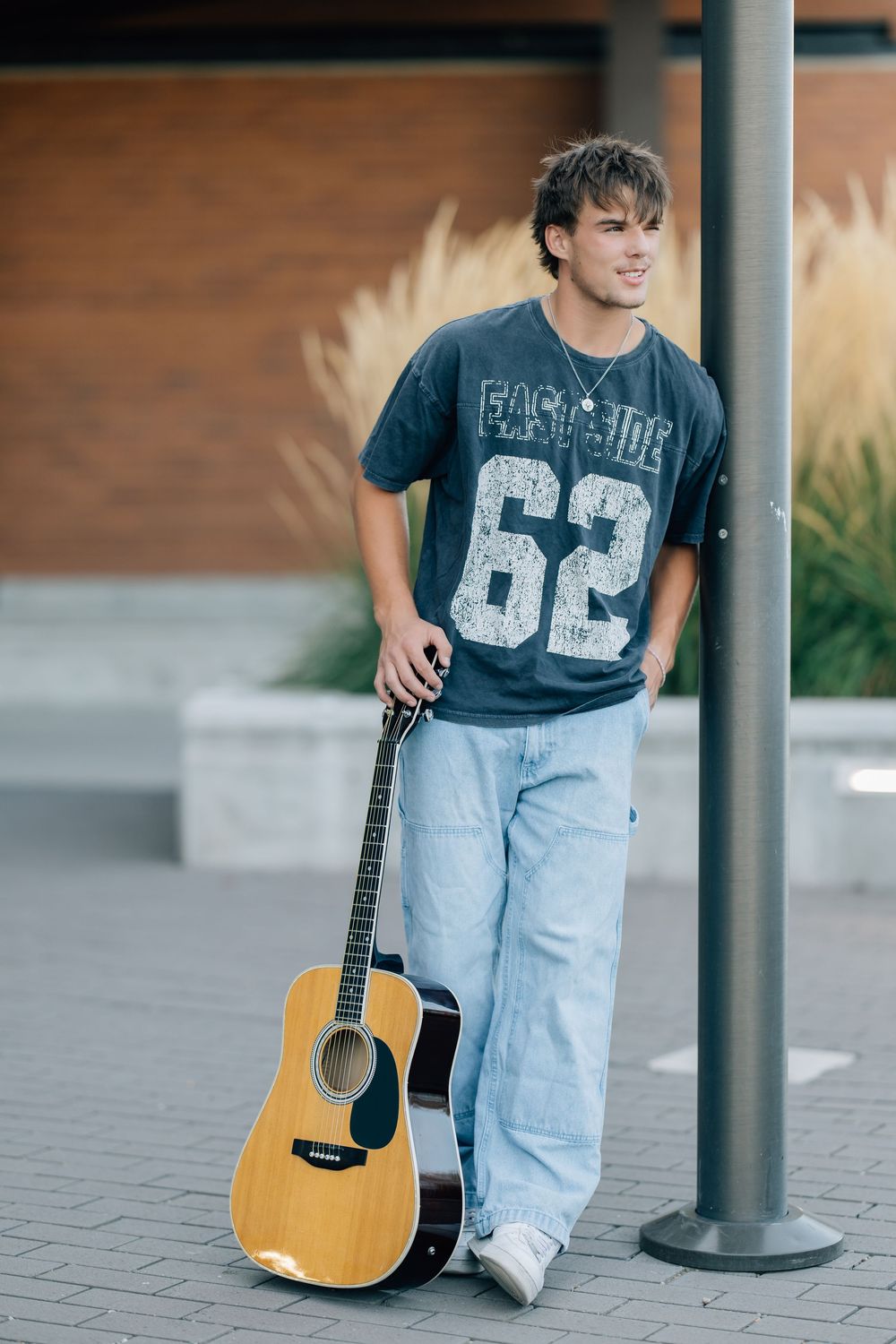 Young man in a grey number 62 t-shirt and jeans leaning on a pole holding an acoustic guitar outdoors.