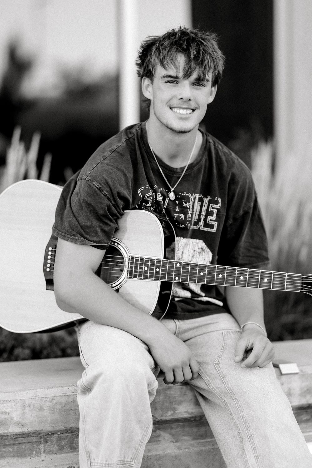Smiling young man holding an acoustic guitar, wearing a graphic tee and jeans, black and white portrait photo.