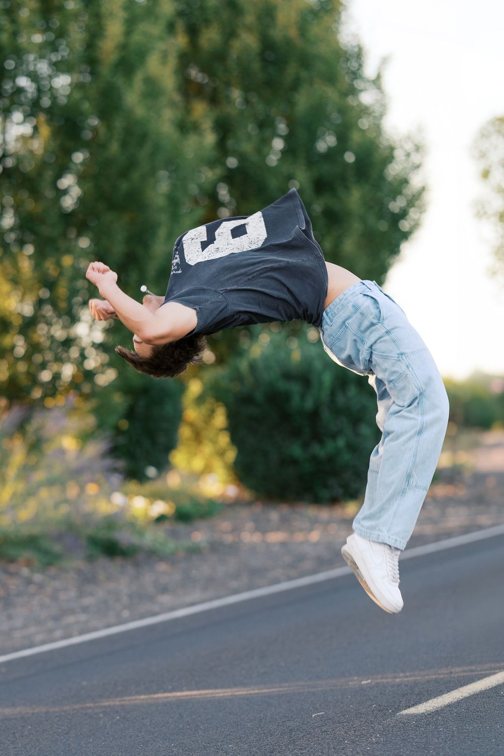 Young person performing a backflip on a road wearing a black jersey and light blue jeans with white sneakers.