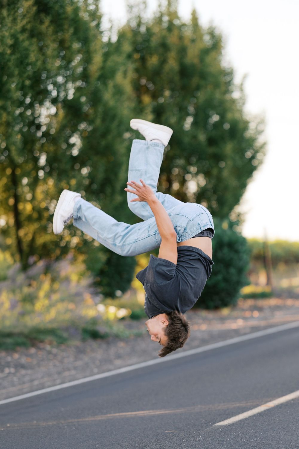 Young man performing a backflip on a road surrounded by lush green trees in natural daylight.