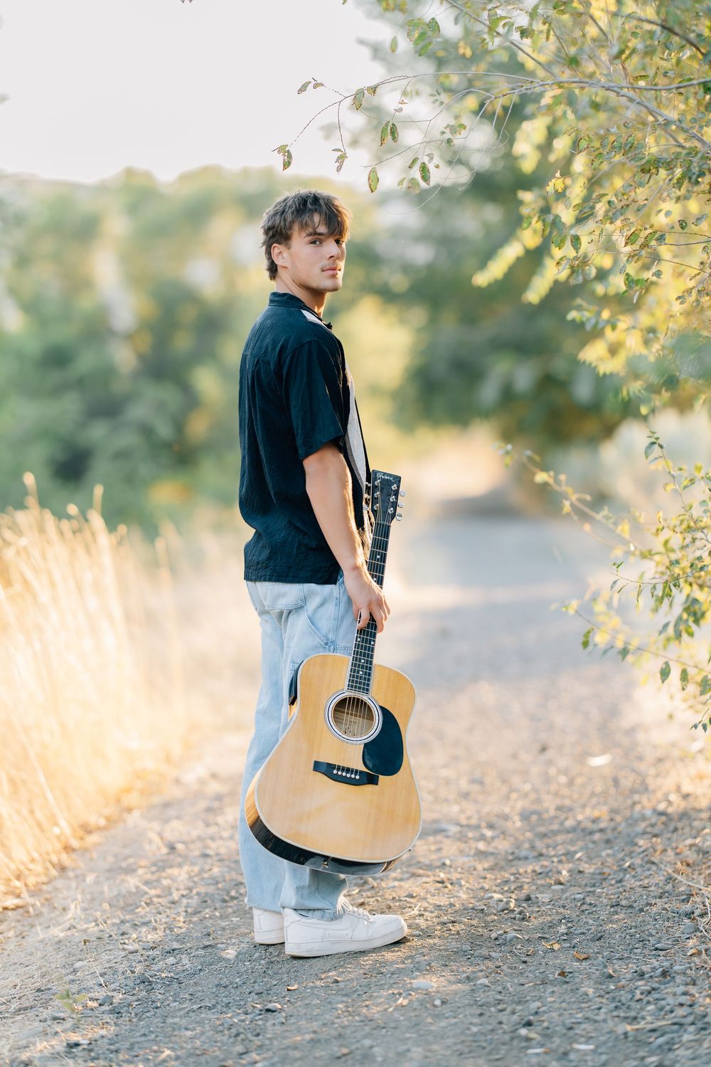 Young man holding an acoustic guitar standing on a rural dirt path surrounded by golden grass and trees.