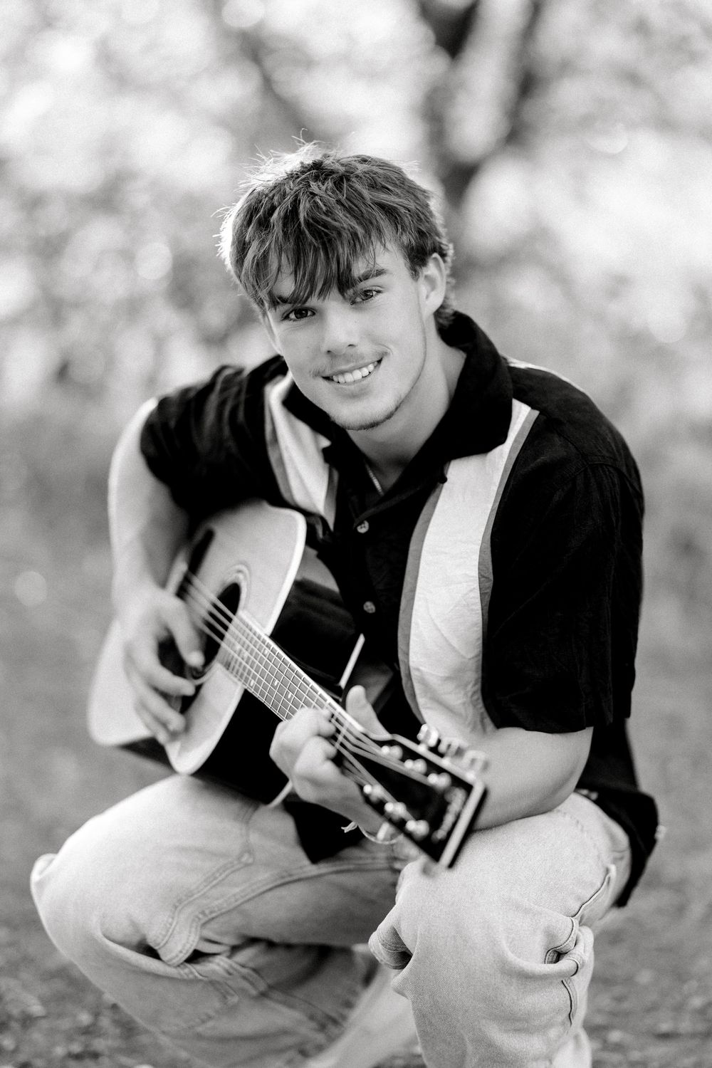 Young man smiling while playing acoustic guitar outdoors, crouching in black-and-white portrait photo.