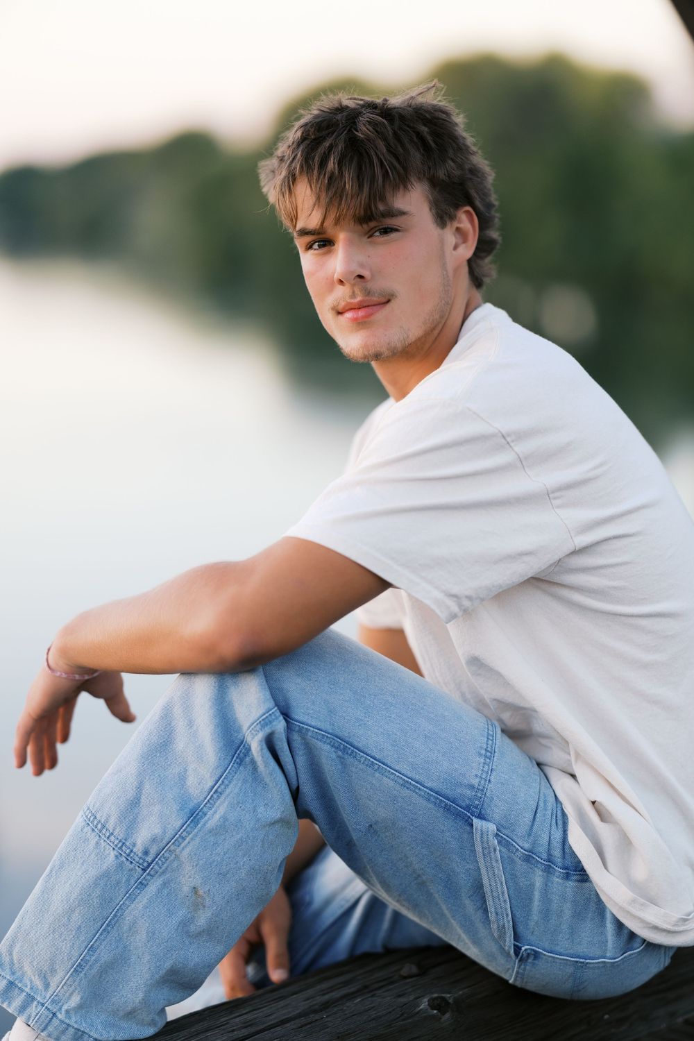Young man in white t-shirt and jeans sitting relaxed by a calm lake with green trees in background.