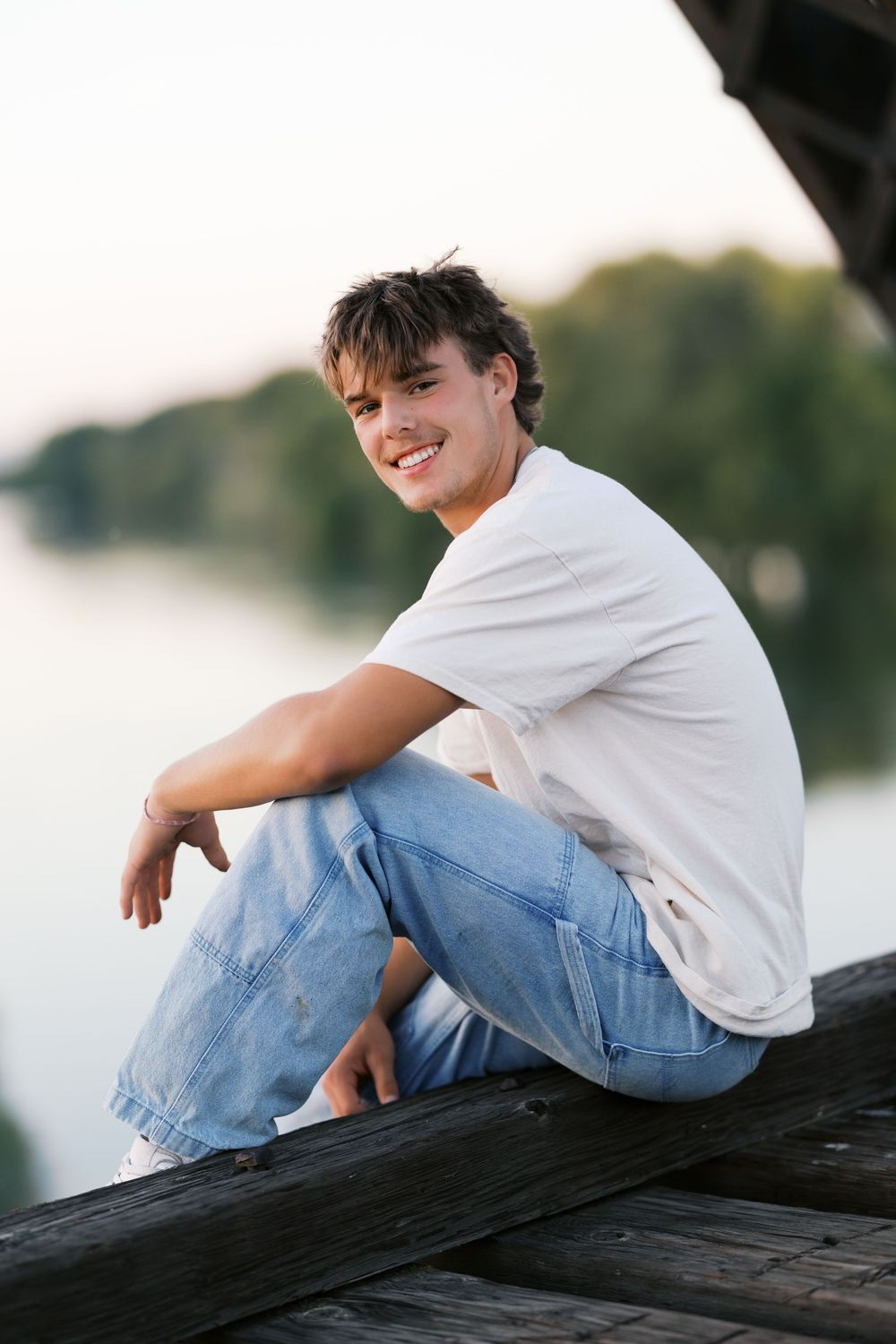 Smiling teenage boy in white t-shirt and jeans sitting on wooden dock by a calm lake at sunset.