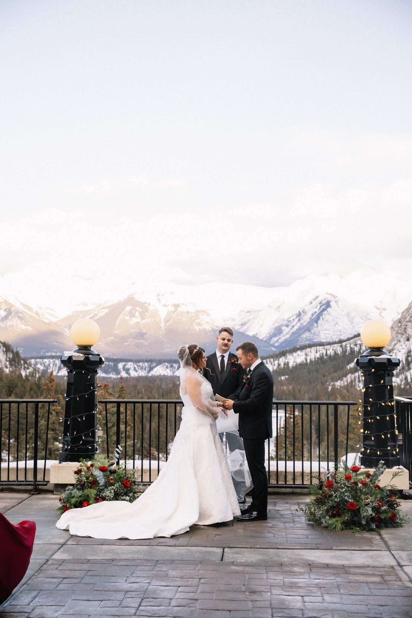 Bride and groom hold hands before their officiant on a Fairmont Banff Springs terrace with snowy peaks behind