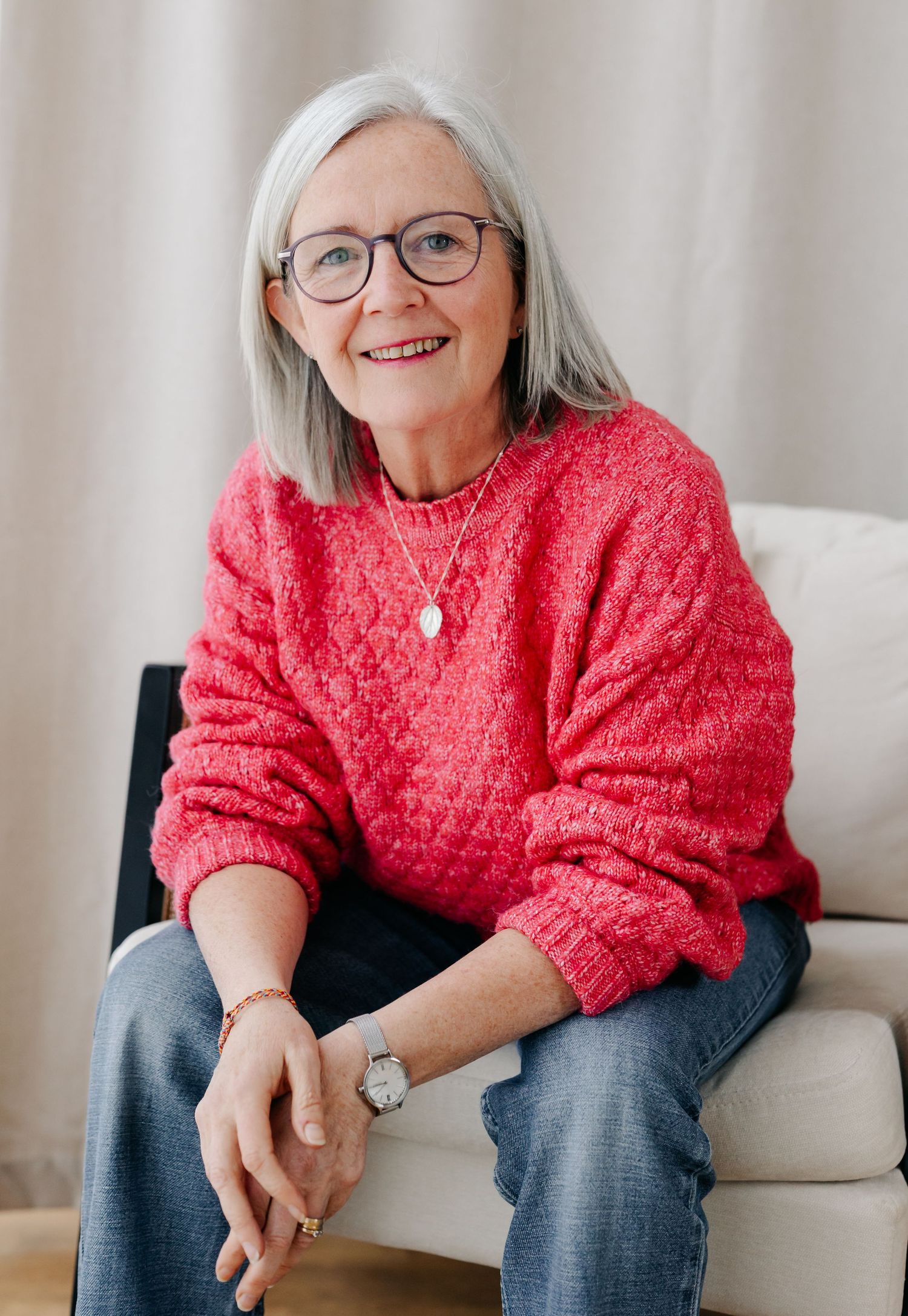 Smiling older woman with gray hair and glasses wearing a red knit sweater, sitting relaxed in a white chair.