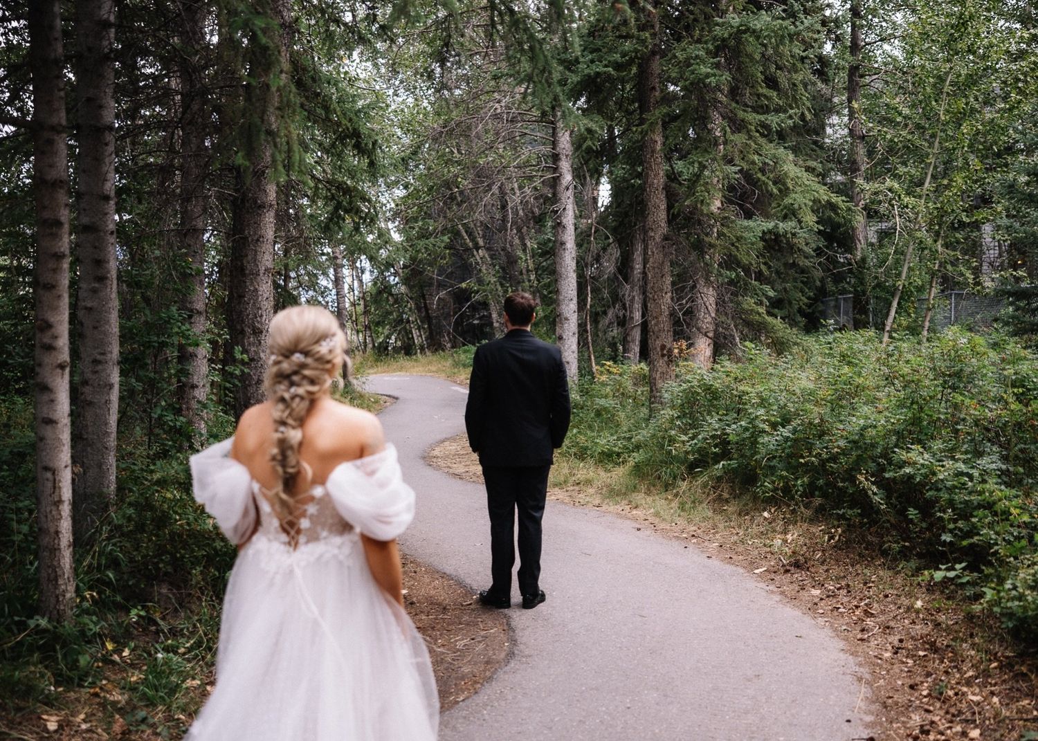 Forest first look on a paved path at the Canmore Nordic Centre, bride walking up behind the groom