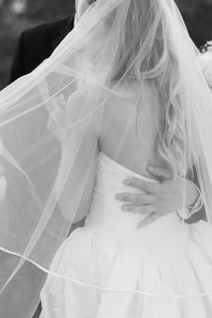 Black and white photo of bride in strapless gown with flowing veil, groom's hand on her waist.