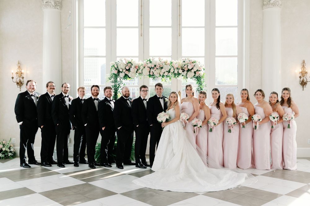 Wedding party in elegant ballroom, groomsmen in black tuxedos and bridesmaids in blush pink gowns.