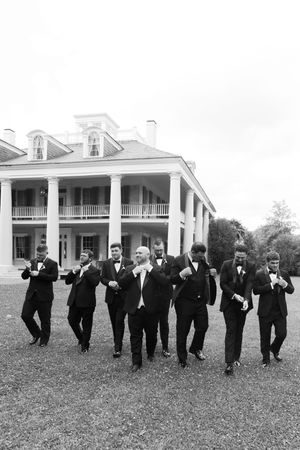 Groomsmen in black tuxedos walking confidently in front of a grand Southern plantation home.
