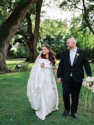 Bride and groom holding hands walking through lush green garden beneath large oak trees.