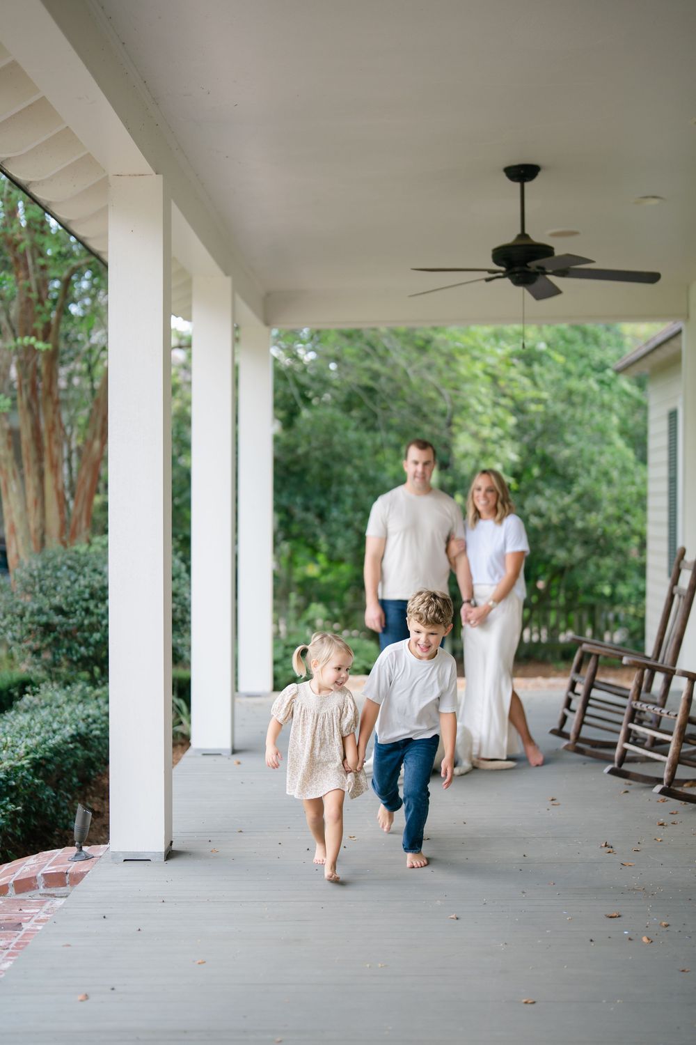 Parents watch two young barefoot children run along covered front porch of classic white Southern home surrounded by trees.