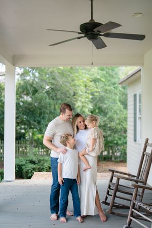 Family of four shares a warm embrace on covered porch with rocking chairs and lush green trees in background.