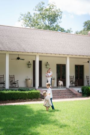Two young children play in green yard in front of classic white Southern farmhouse with covered porch and rocking chairs.