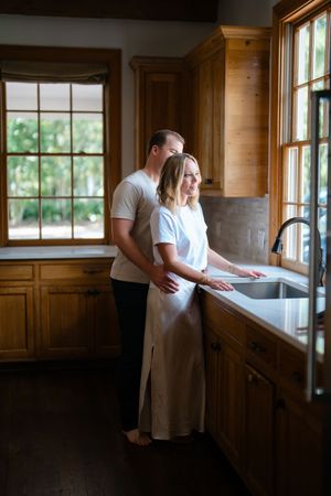 Couple shares a tender moment together in a warm wooden kitchen with natural light streaming through large windows.