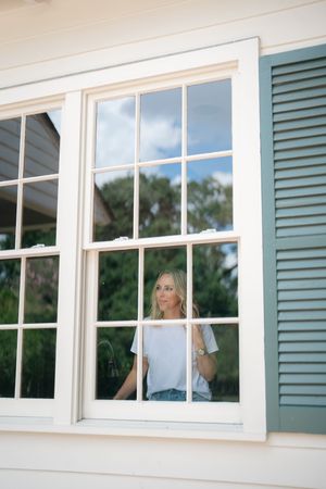 Blonde woman gazing through a white-framed window with blue shutters on a sunny day.