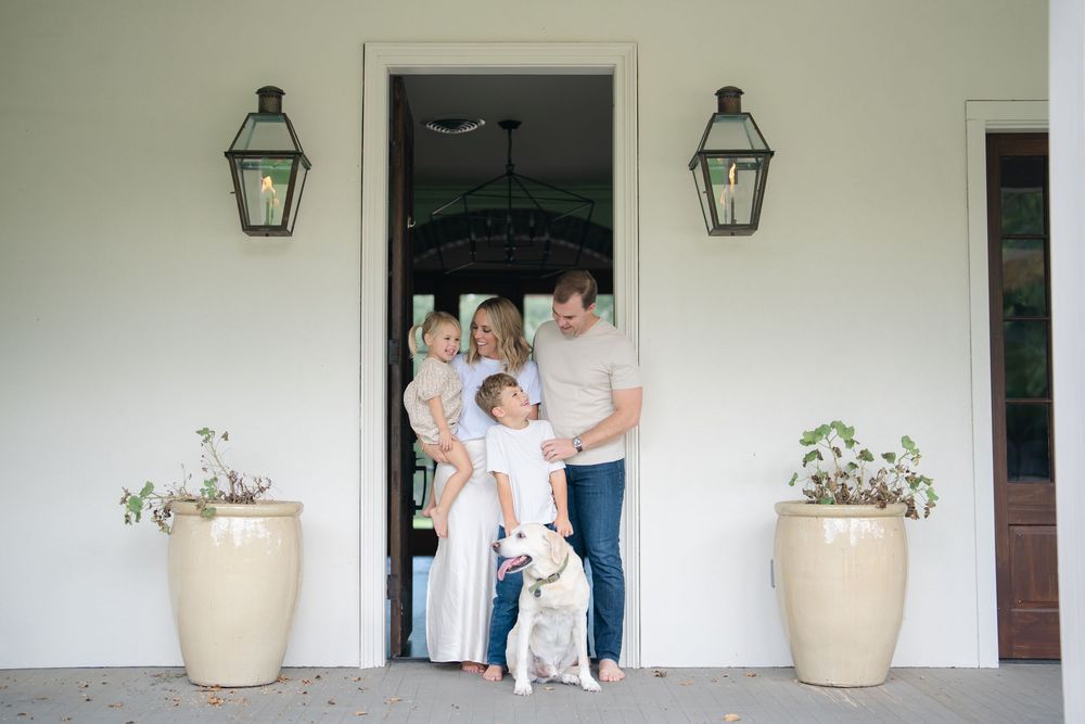 Happy family with two young children and white dog pose in doorway of white Southern home with lanterns.