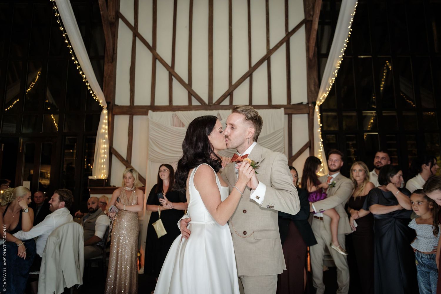 Bride and groom share a kiss on the dance floor surrounded by wedding guests in a rustic barn venue.