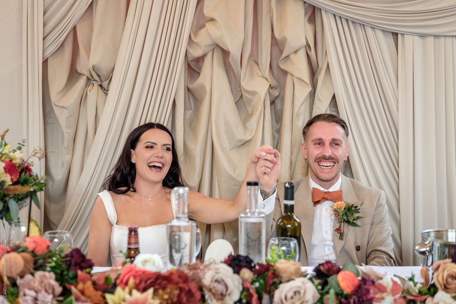 Happy couple laughing and raising glasses at wedding reception table adorned with colorful floral arrangements.