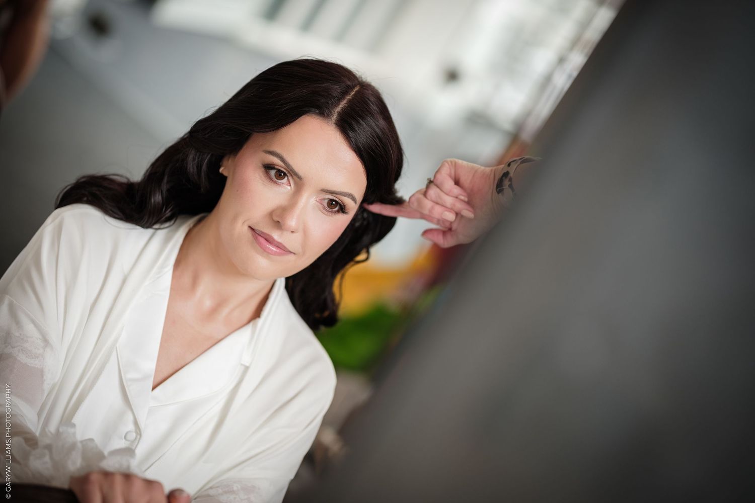 A young brunette woman in a white blouse sits indoors, smiling softly while someone points near her face.