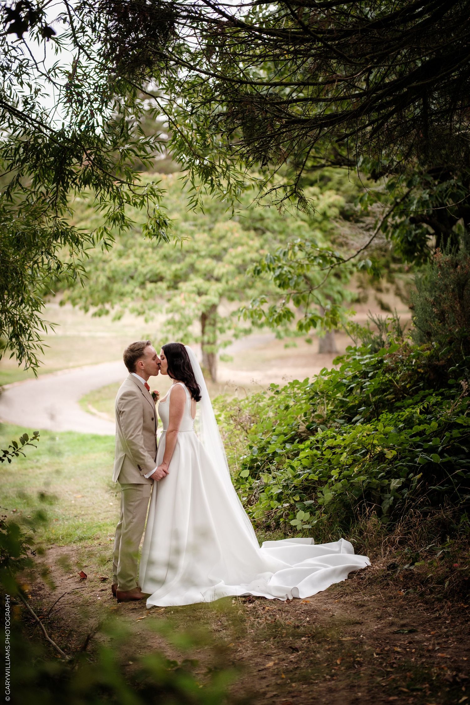 A bride and groom share a romantic kiss under lush green trees during an outdoor wedding ceremony.