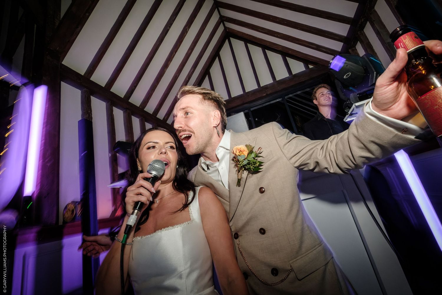 Bride and groom singing joyfully at wedding reception party with microphone and purple lights.