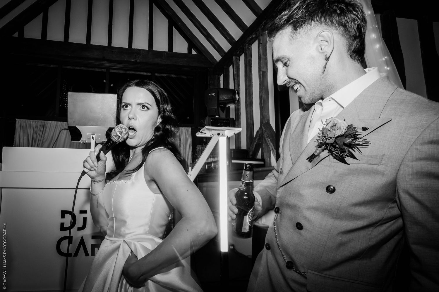 Bride singing into microphone with groom at wedding reception in black and white.