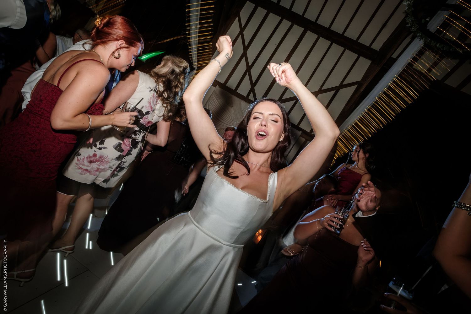 Bride in white dress dancing joyfully at wedding reception with guests celebrating in background.