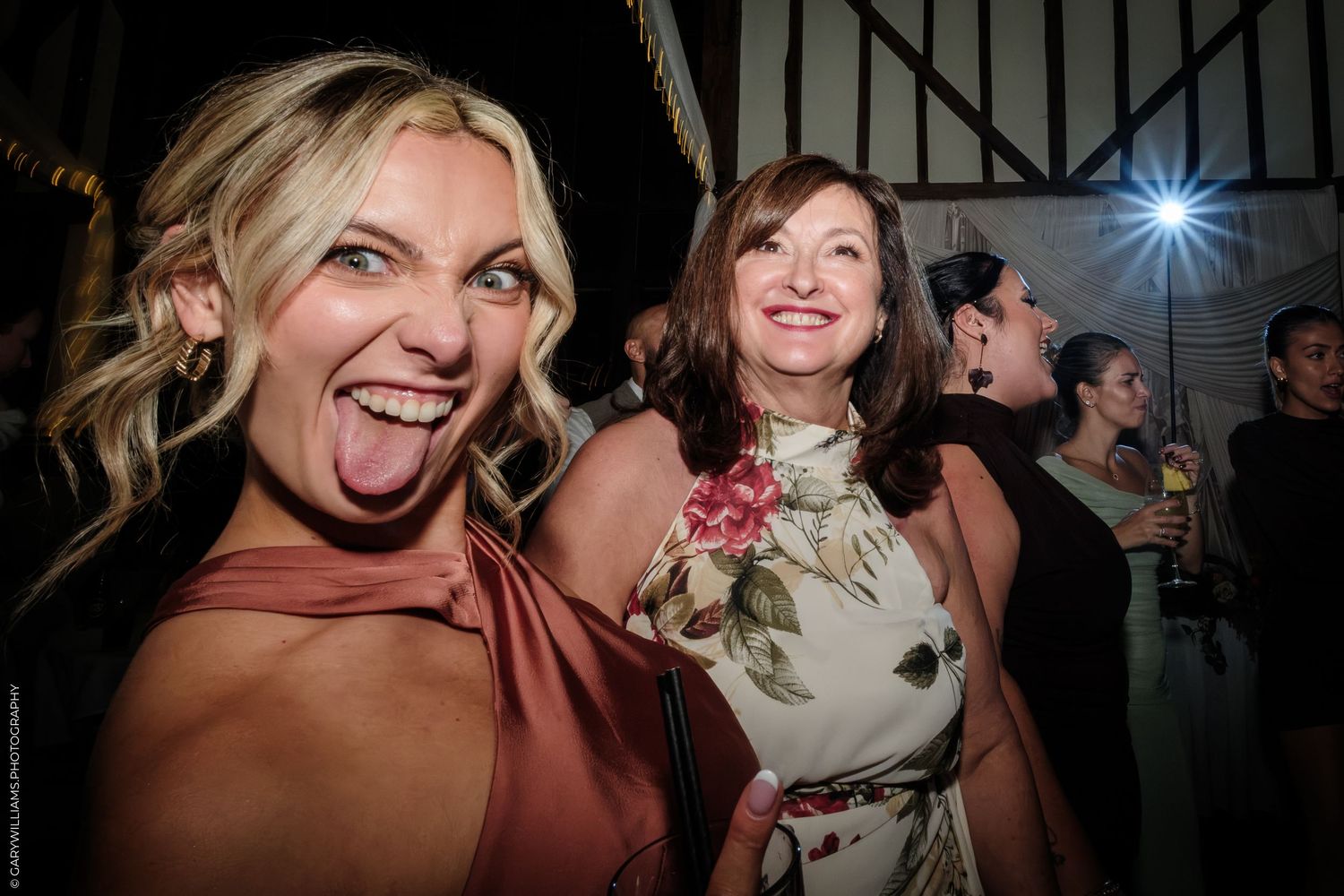 Two women smiling and laughing at a wedding reception, one playfully sticking out her tongue.