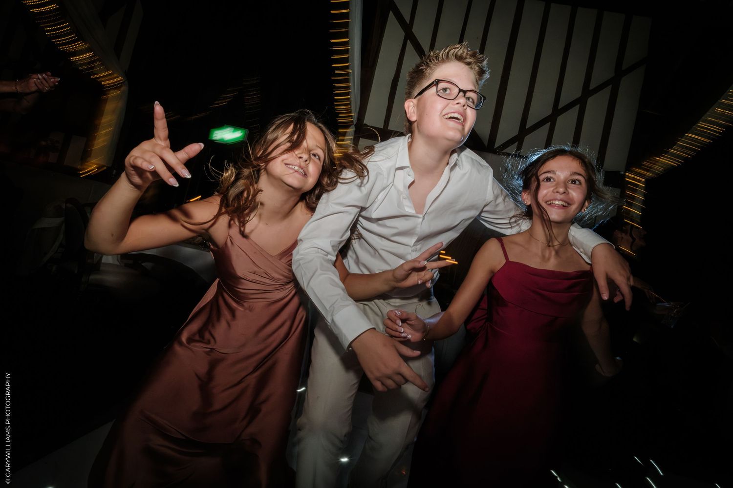 Three children dancing joyfully at a wedding reception, wearing formal attire in pink, white, and red.