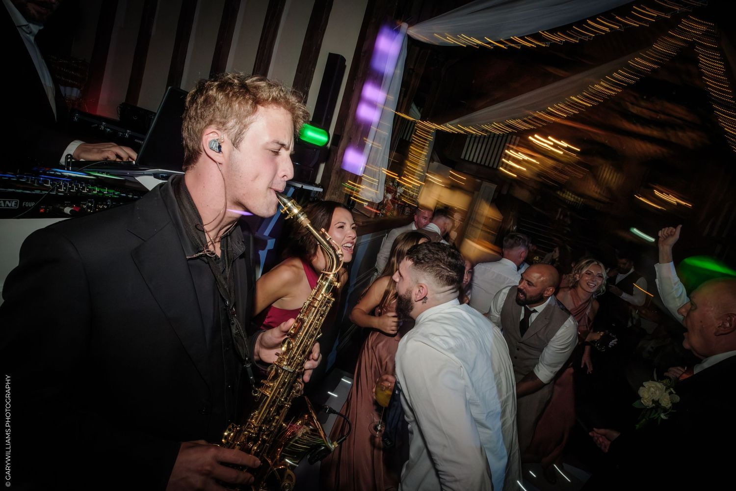 A saxophonist in a black suit performs energetically at a lively wedding reception with guests dancing in the background.