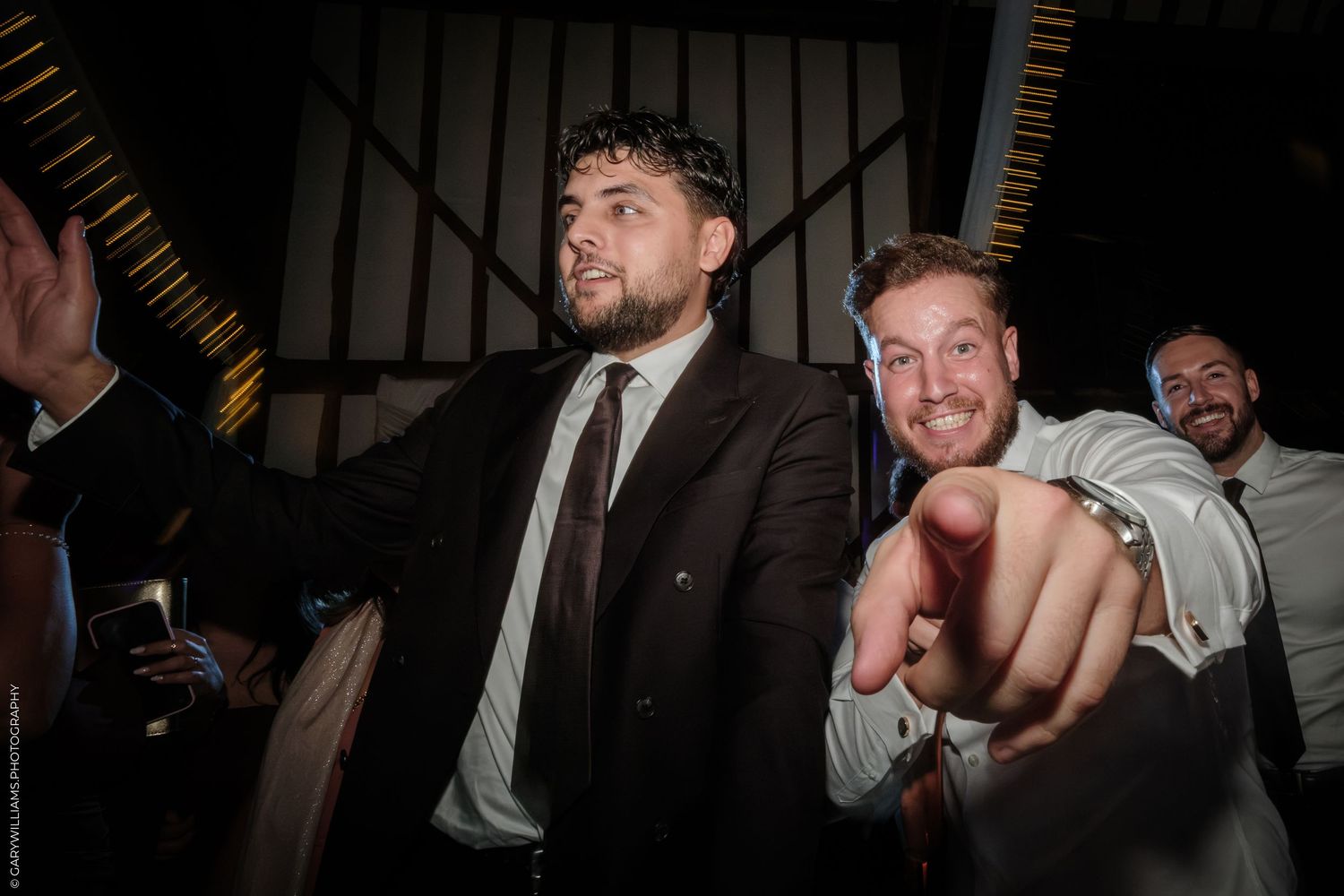 Two men in suits smiling and pointing at camera during a formal event or wedding reception.