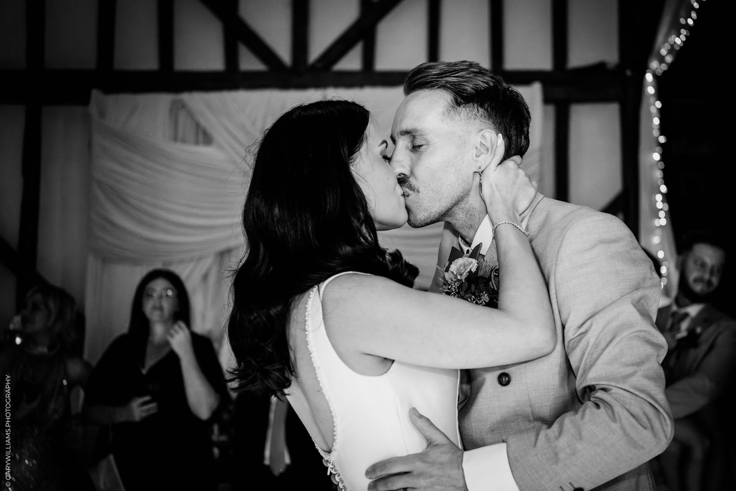 Black and white photo of a couple sharing their first kiss at a wedding reception with guests watching in the background.