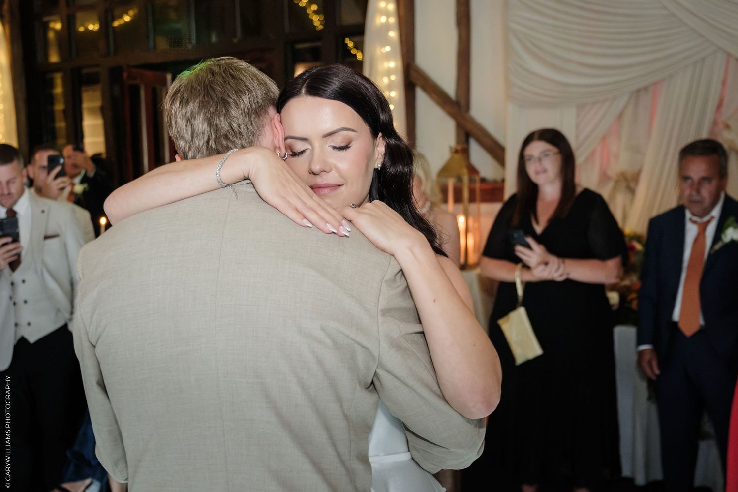Bride and groom share an emotional first dance embrace at elegant wedding reception with guests watching in background.