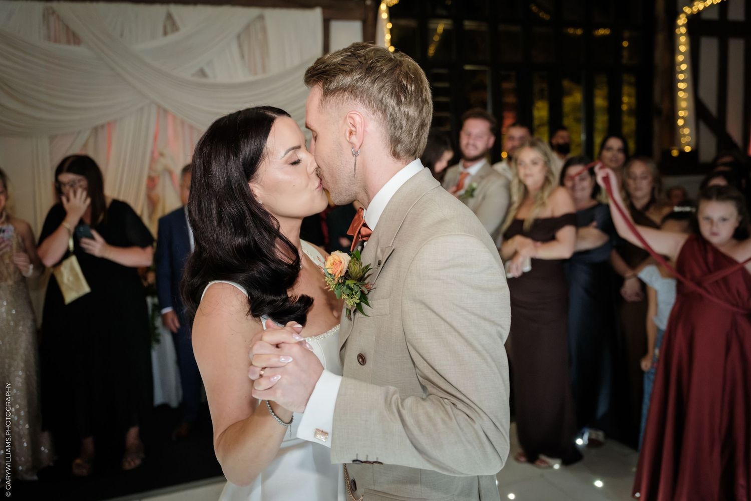 Bride and groom share their first dance kiss at wedding reception with guests celebrating in background.