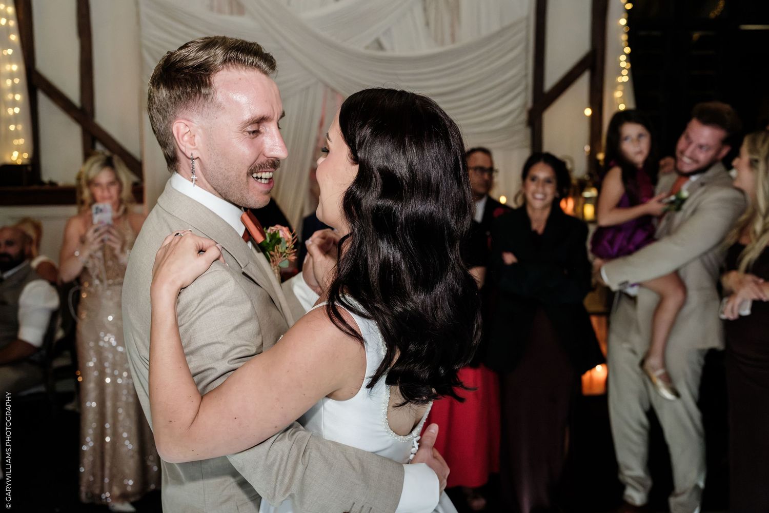 Bride and groom share their first dance at wedding reception with guests smiling in background.