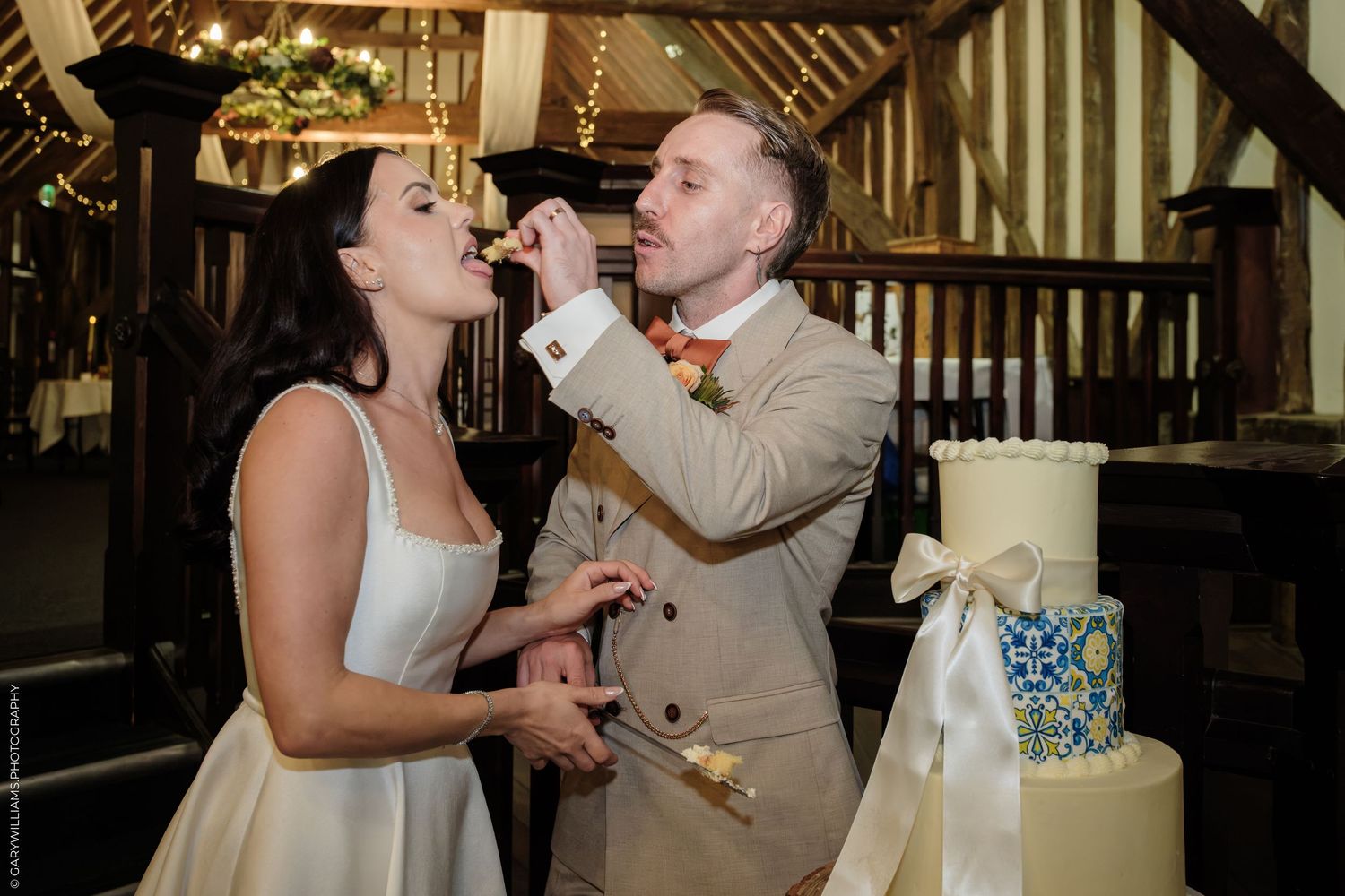 Bride and groom cutting wedding cake in rustic barn venue, groom feeding cake to bride in elegant white dress.