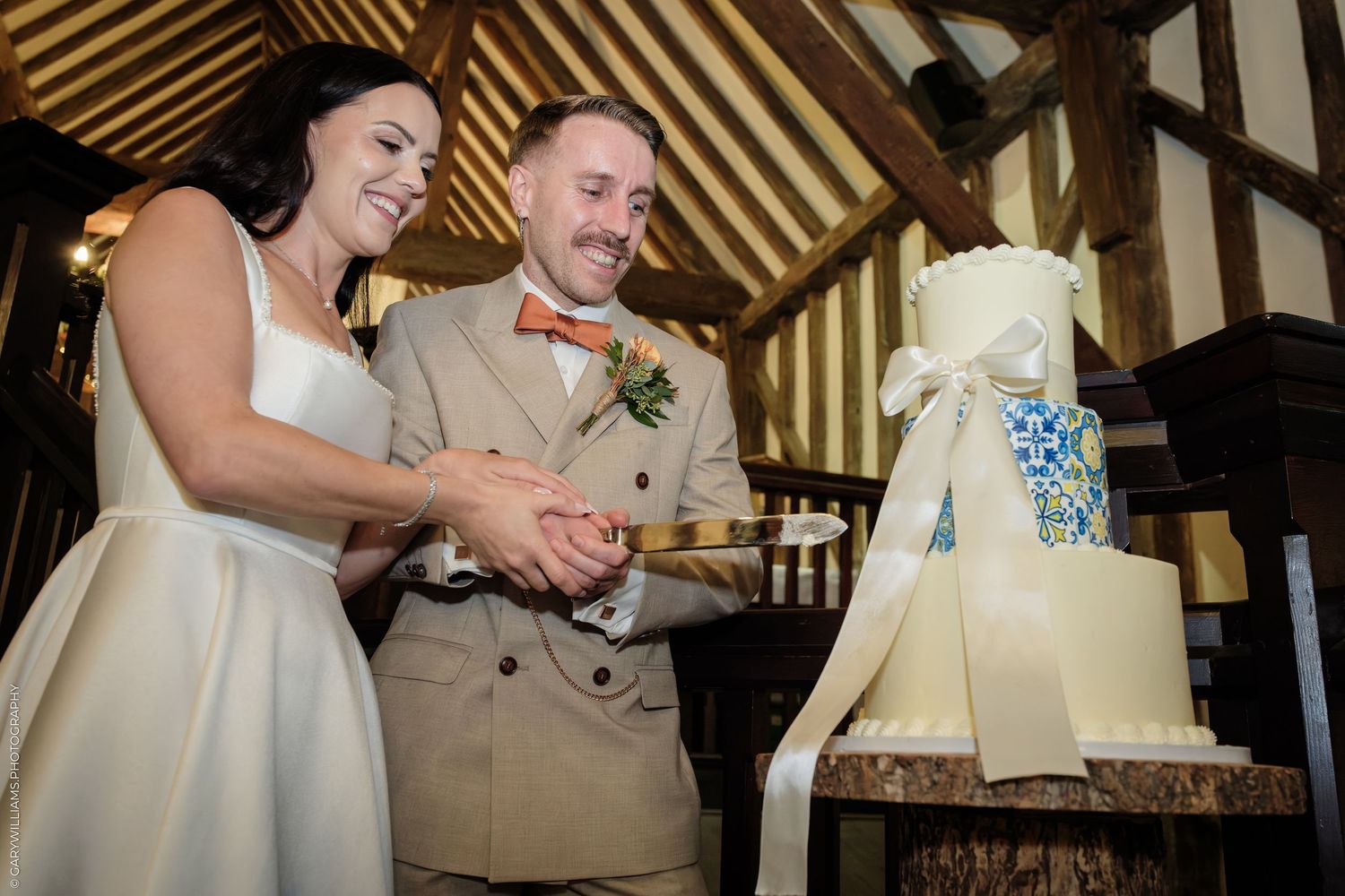 Bride and groom smiling while cutting their elegant white wedding cake together in a rustic barn venue.