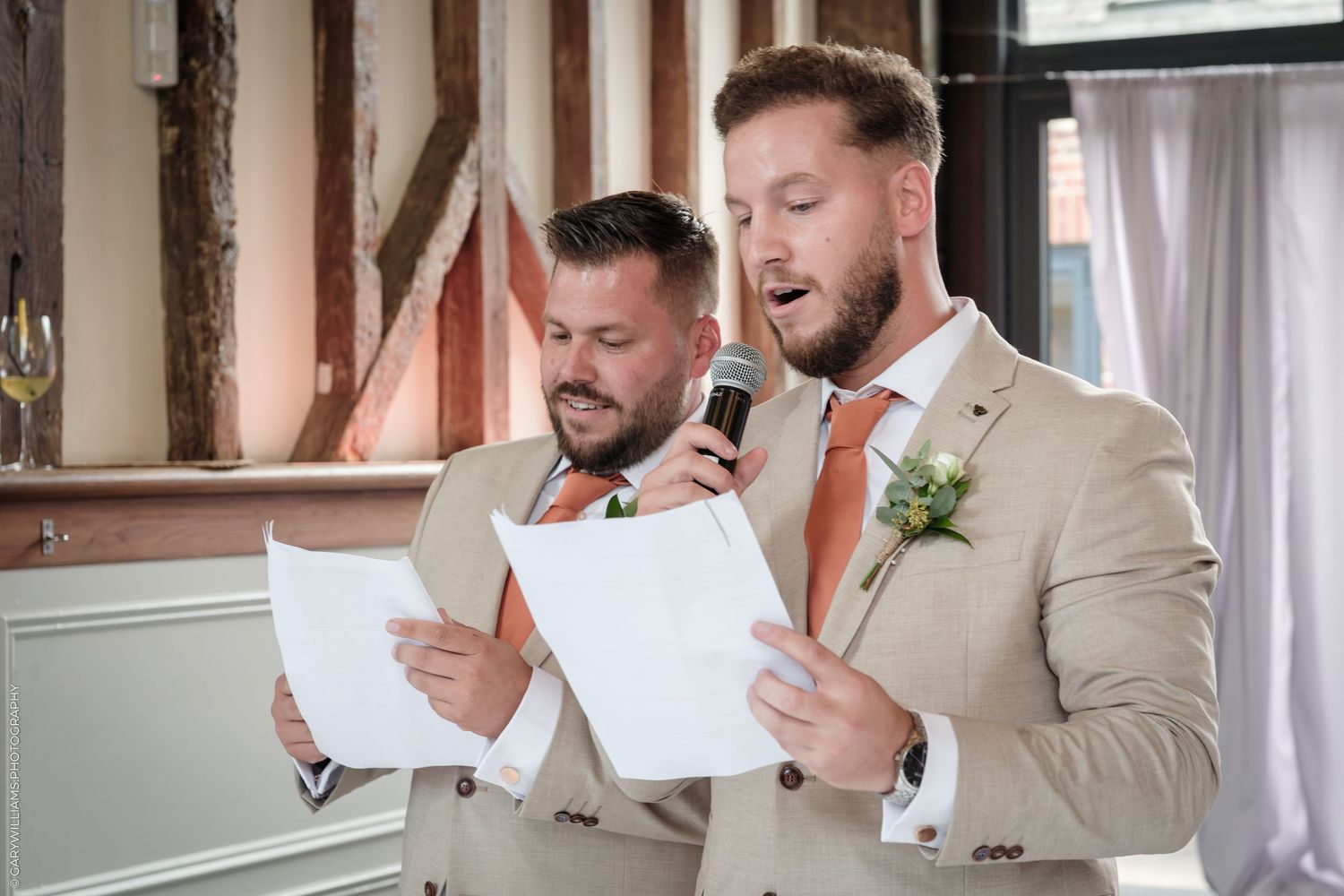 Two groomsmen in tan suits with orange ties delivering a wedding speech together at an indoor venue.
