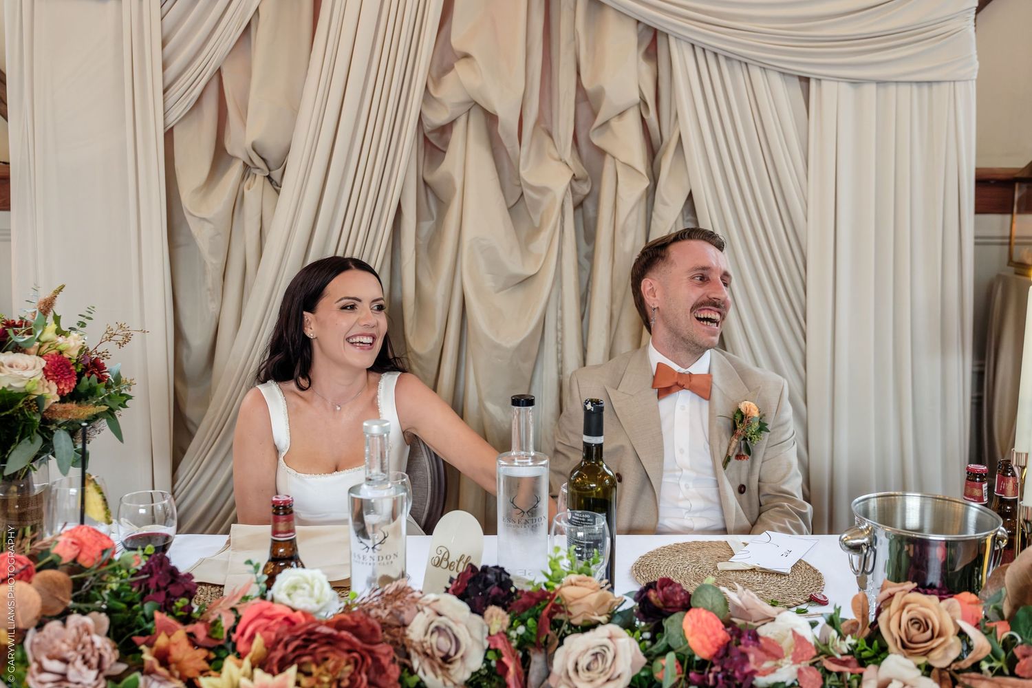 Bride and groom laughing joyfully at wedding reception head table adorned with lush floral arrangements.
